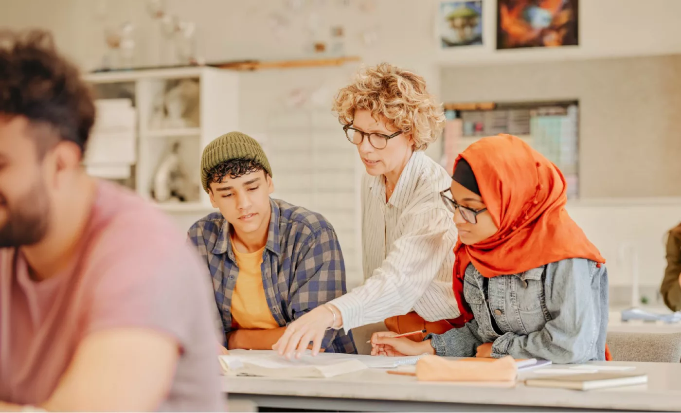 A female teacher with glasses leans over a desk to help two students—a young woman in an orange hijab and a young man in a beanie—in a classroom setting.